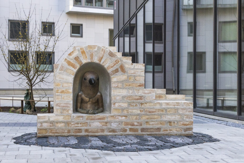 Ett foto av ett konstverk av tegel och brons som står integrerad mellan flerfamiljshus på en gågata.