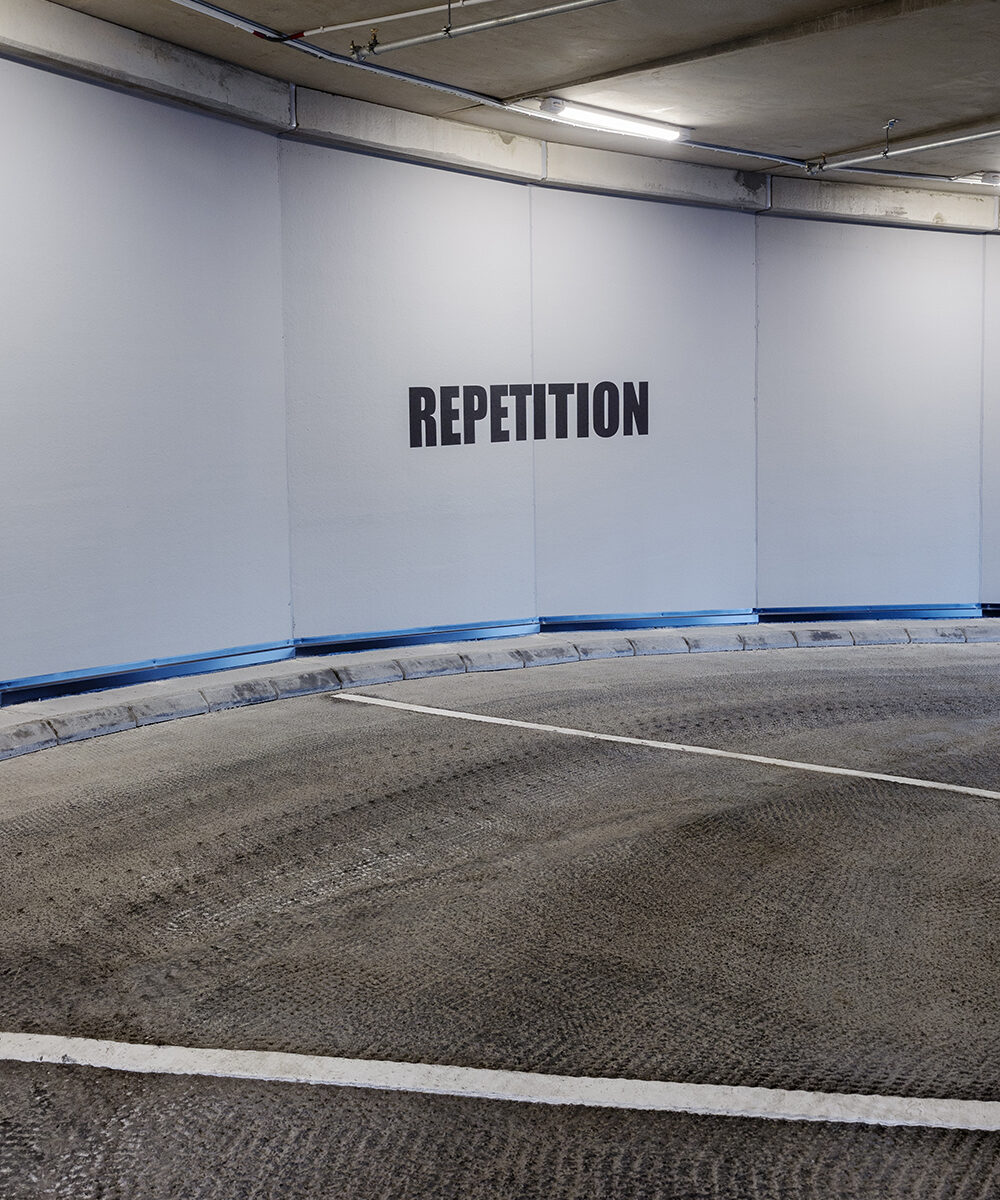 Ordet "REPETITION" står skrivet på en vit vägg framför vägbanan.