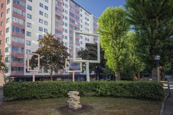 The Common Home En mindre träskulptur av en hög med böcker syns framför en häck. Bakom häcken tornar fönsterskulpturerna upp sig mot de höga husfasaderna.